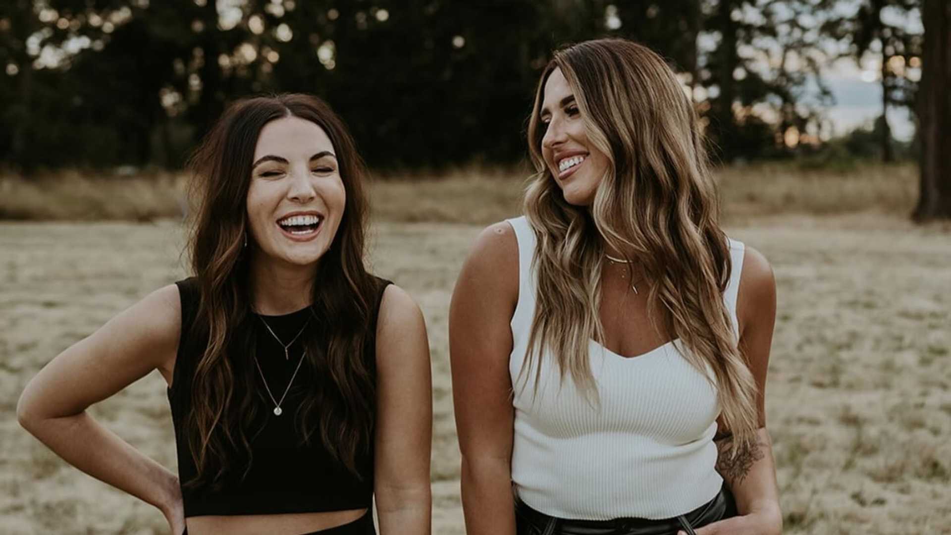 Two women smiling and standing in a field, dressed in casual black and white outfits.