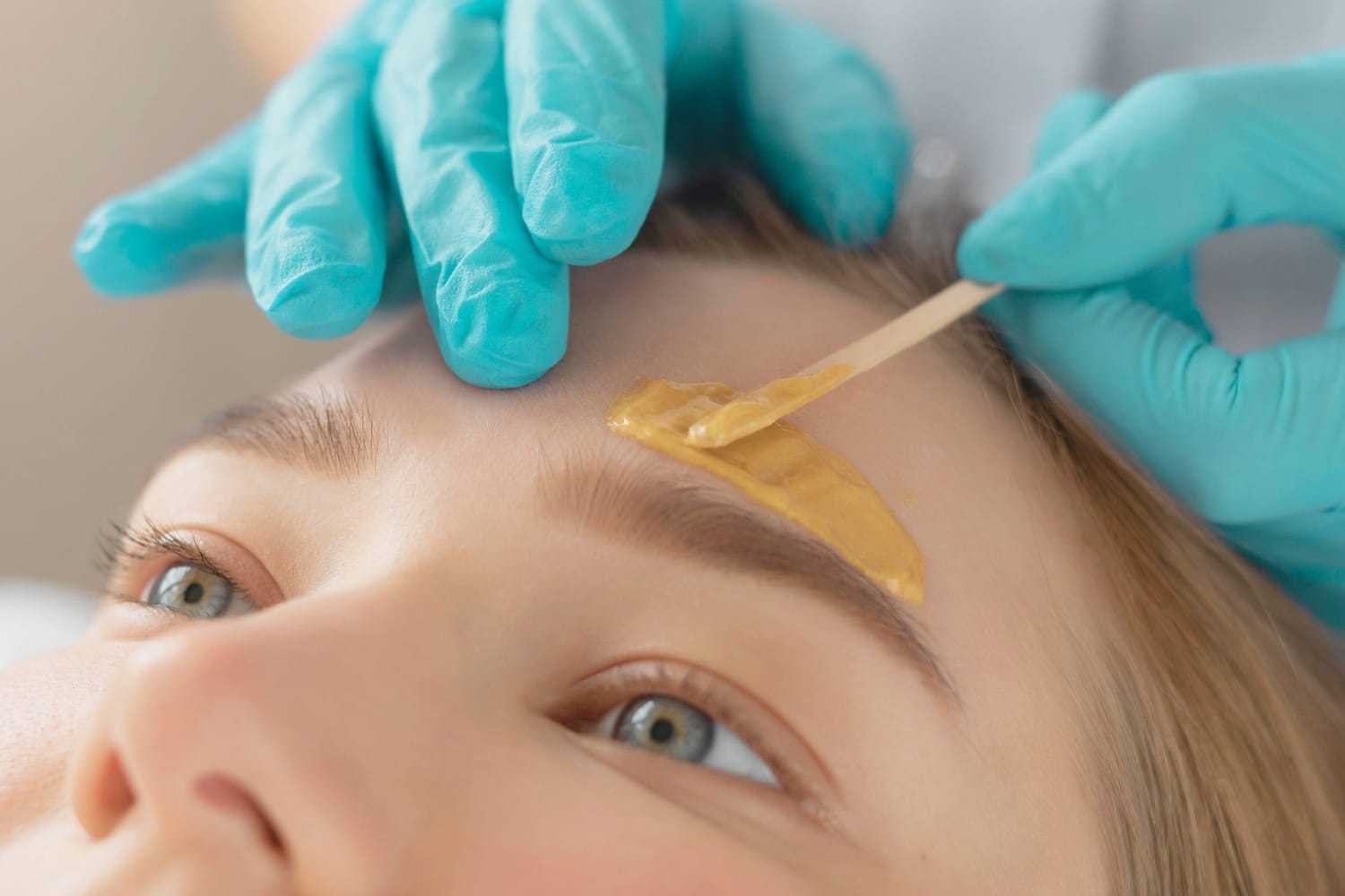 Eyebrow waxing procedure with gloved hands applying wax on a person's eyebrow.