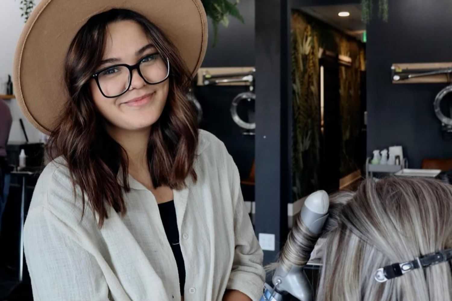 Woman in hat smiling at hair salon, curling client's hair with a wand.
